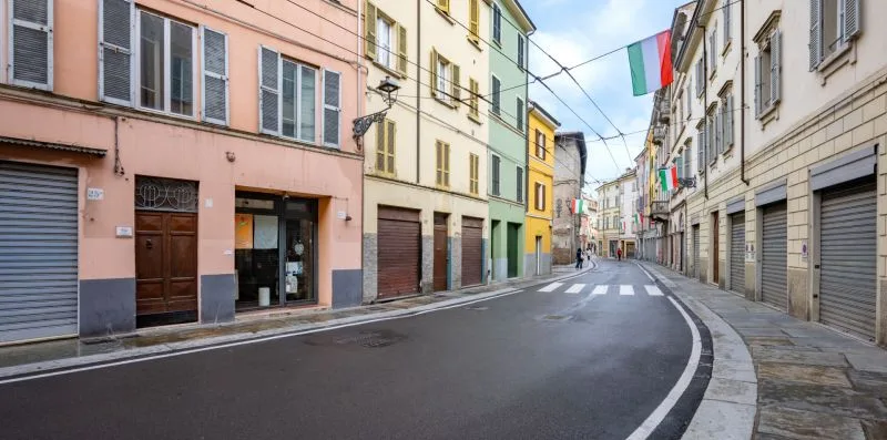 Una calle urbana estrecha se curva entre edificios de colores pastel con banderas italianas.