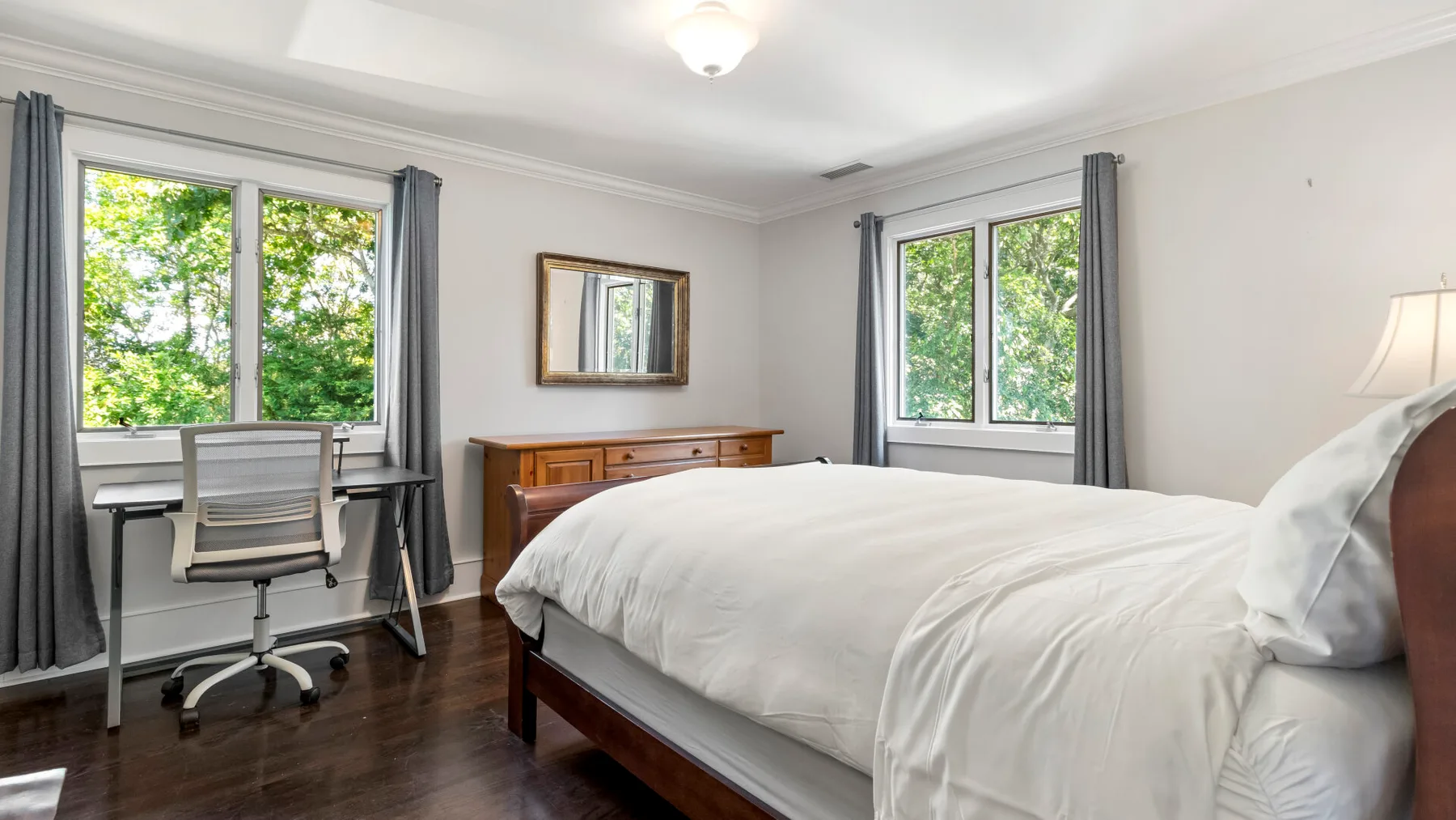 Un dormitorio con una cama, un escritorio y ventanas con vista a árboles frondosos.