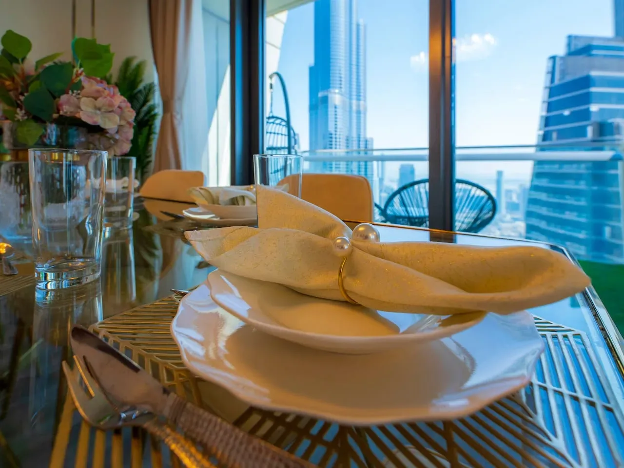 A dining table is set with plates and folded napkins beside large windows.