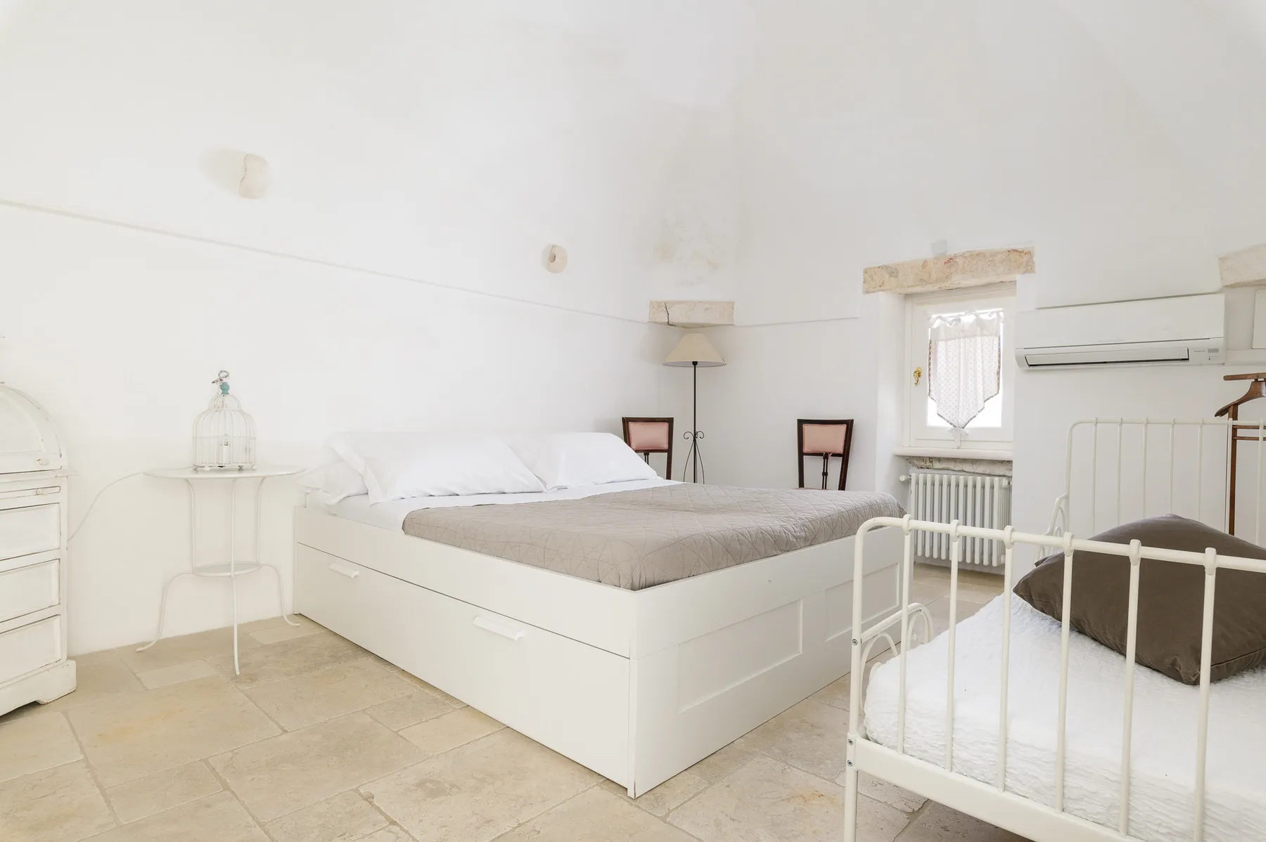 A bright bedroom with a large bed and a white crib beside the window.
