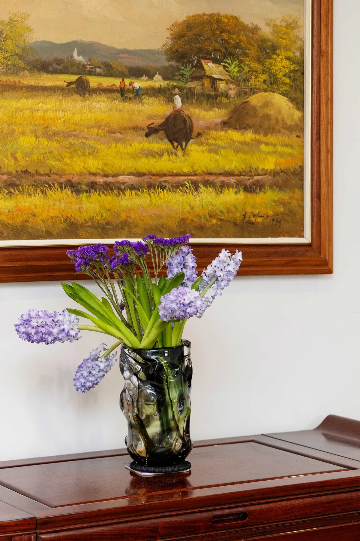 Un jarrón con flores moradas y blancas está sobre una mesa de madera bajo un cuadro enmarcado.