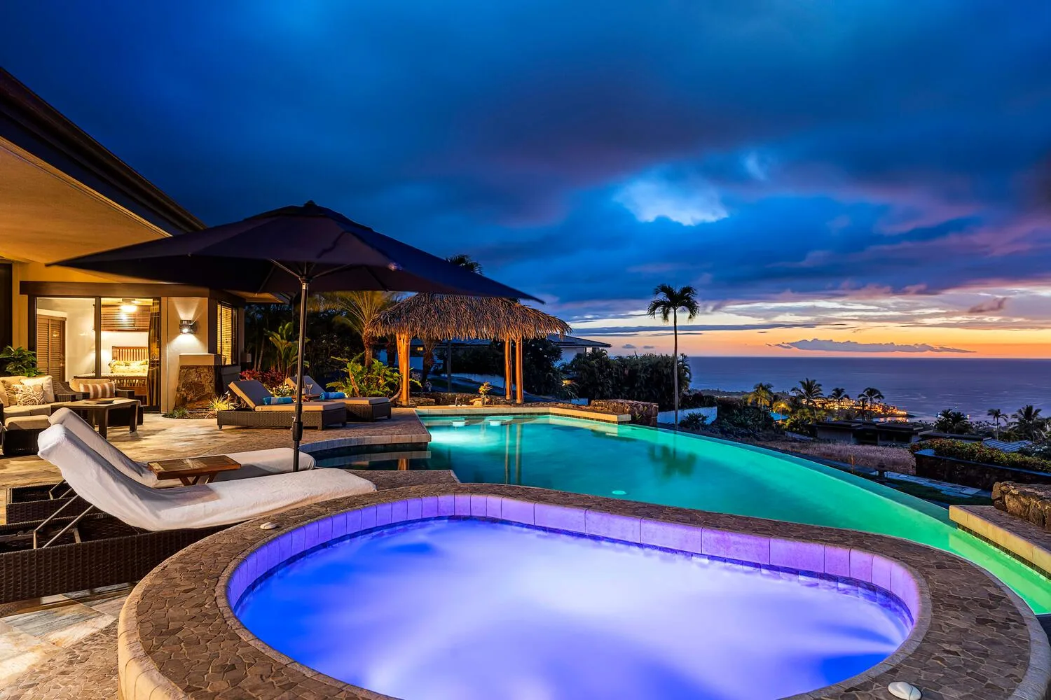 Una piscina al aire libre y un spa iluminado miran al océano bajo un cielo colorido al atardecer.