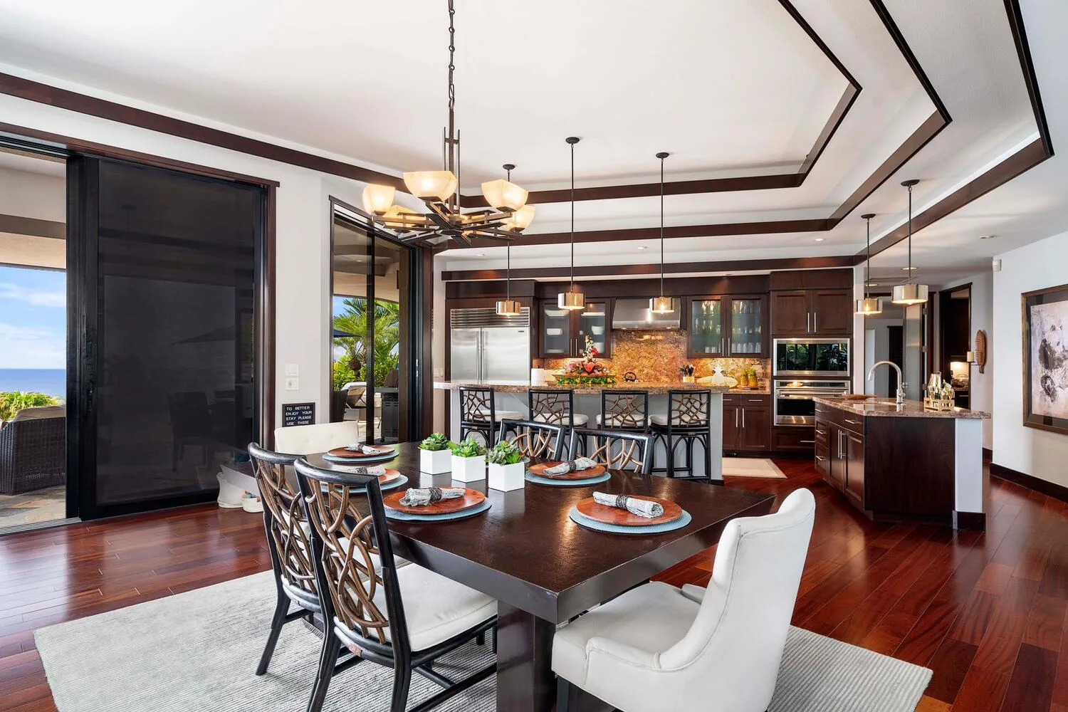 Un comedor se abre a una cocina con gabinetes oscuros y una mesa grande.