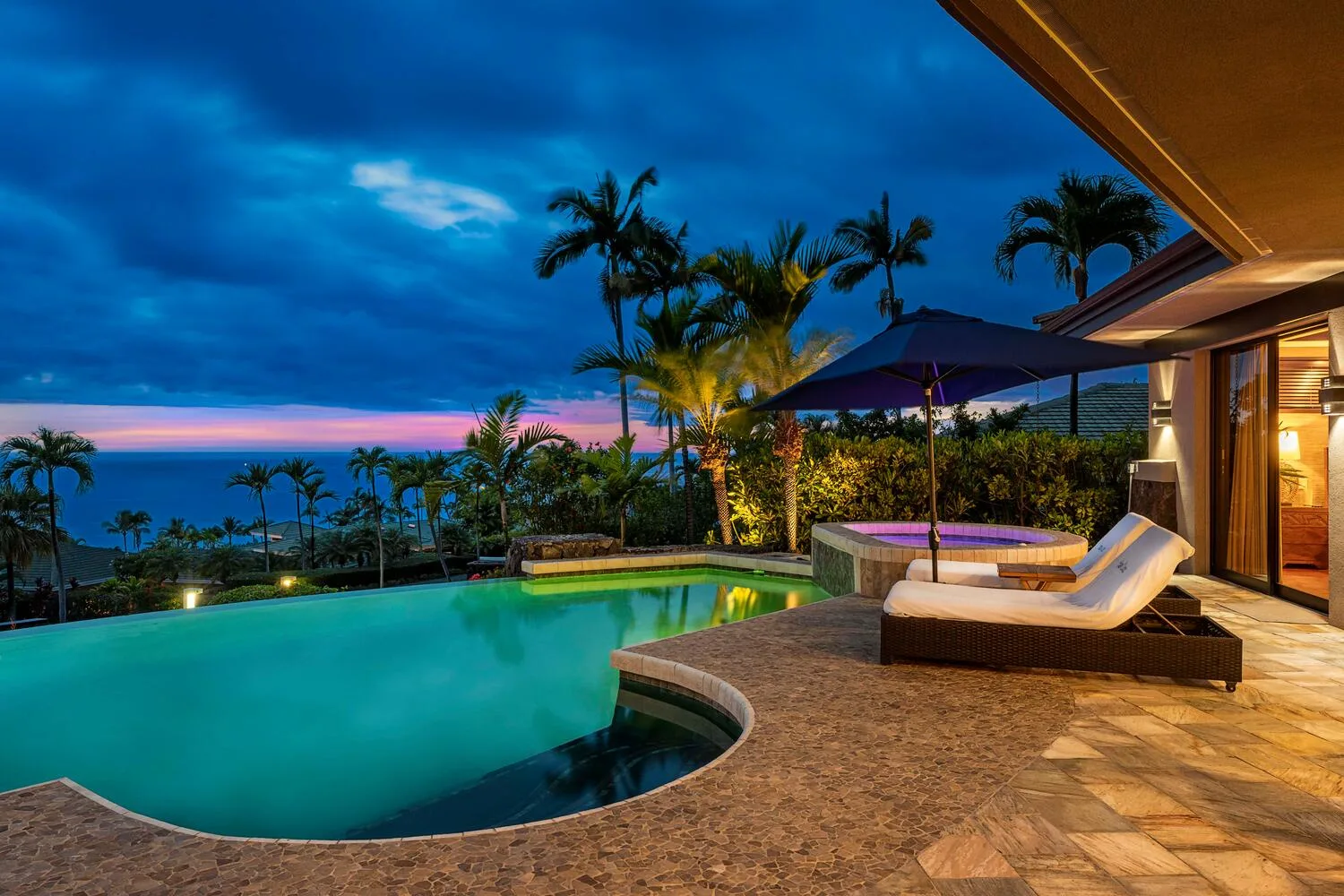 Una piscina exterior junto a un patio con asientos reclinables al anochecer.