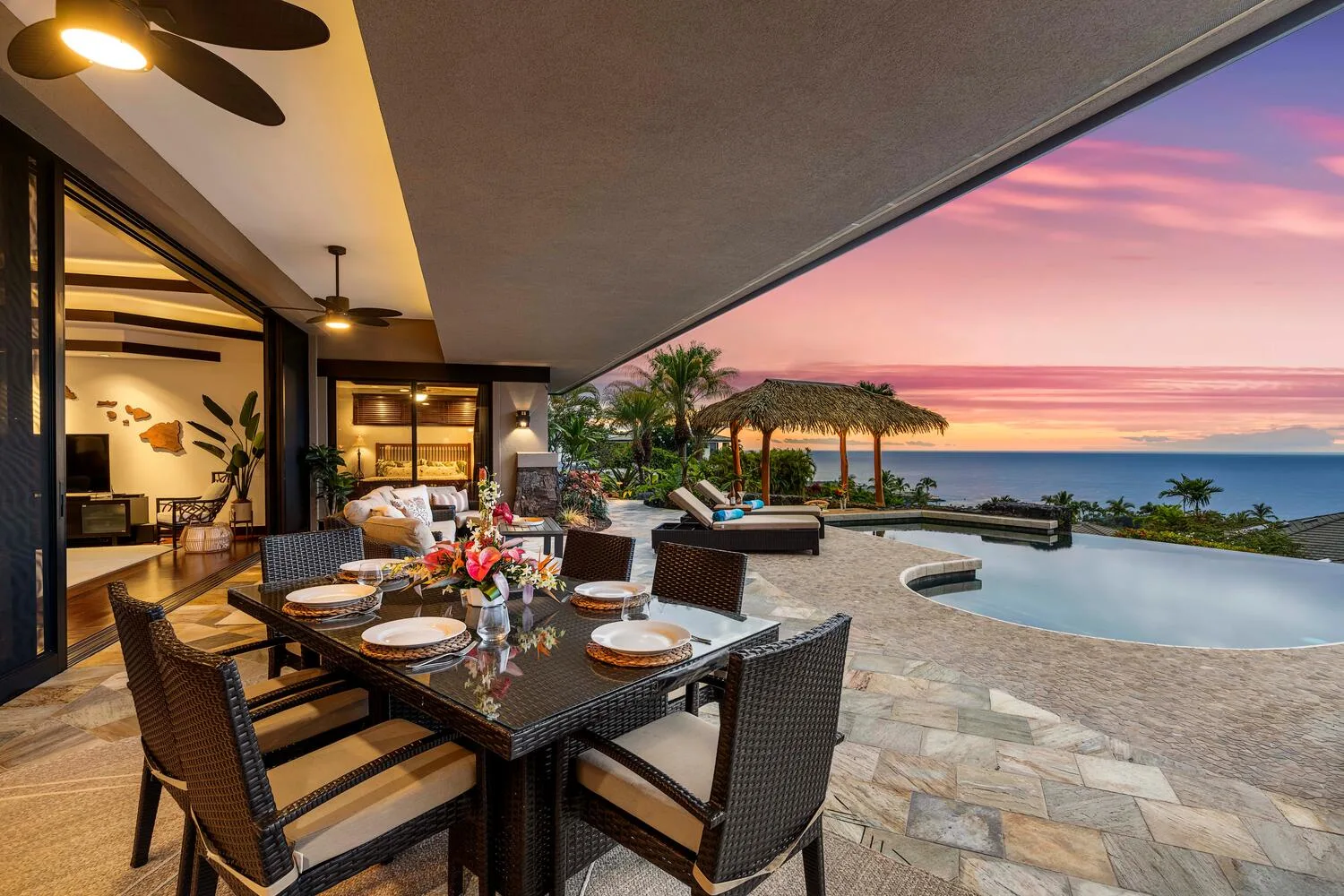 Una zona de comedor exterior cubierta da a una piscina y al mar al atardecer.