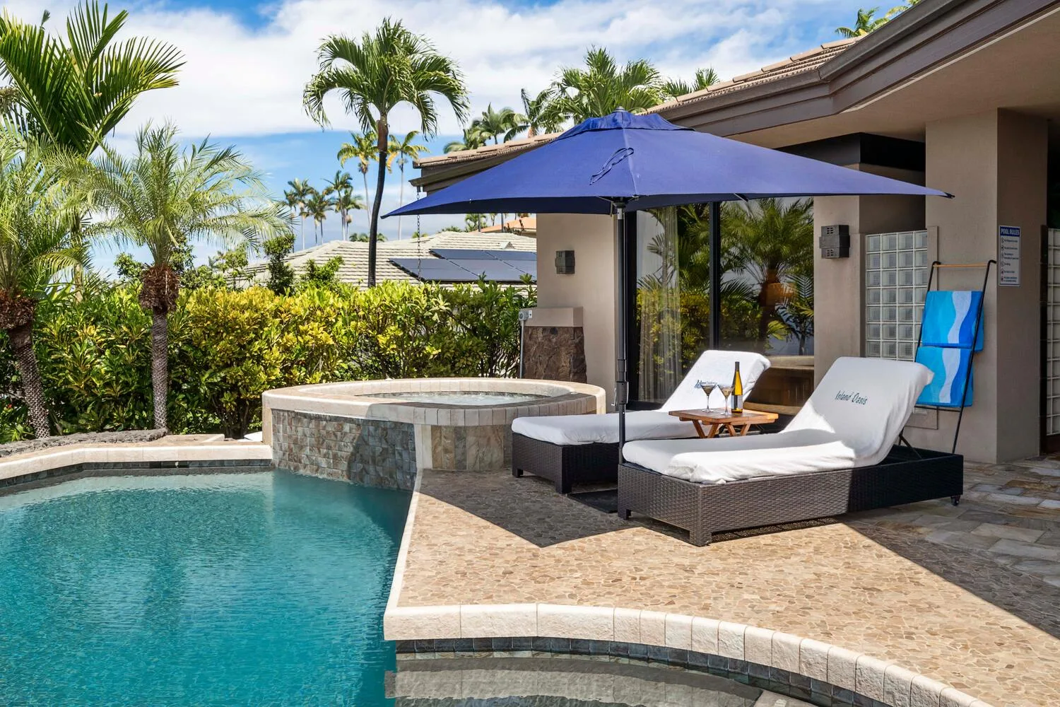 Un patio junto a la piscina tiene asientos acolchados bajo una sombrilla azul.