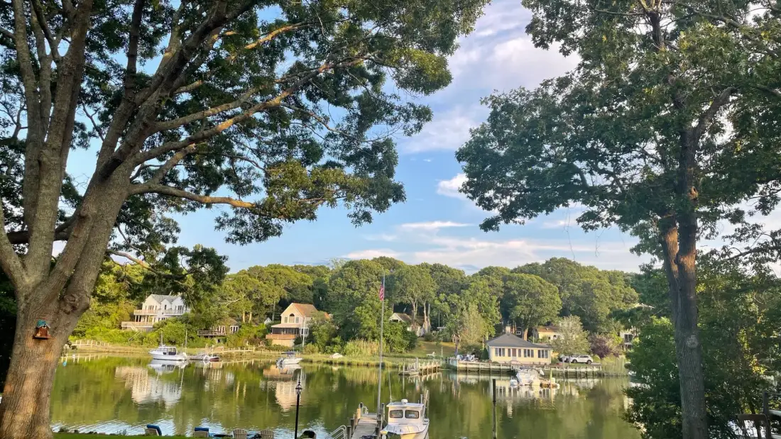 A calm waterfront scene shows homes and boats reflected in the water.