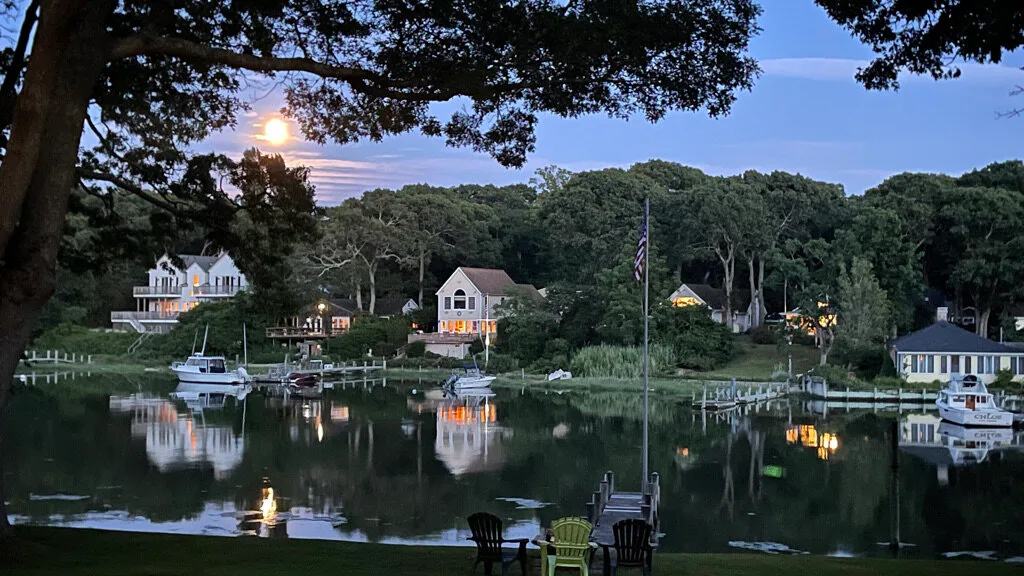 A waterfront scene at dusk, with homes and boats reflected in calm water.