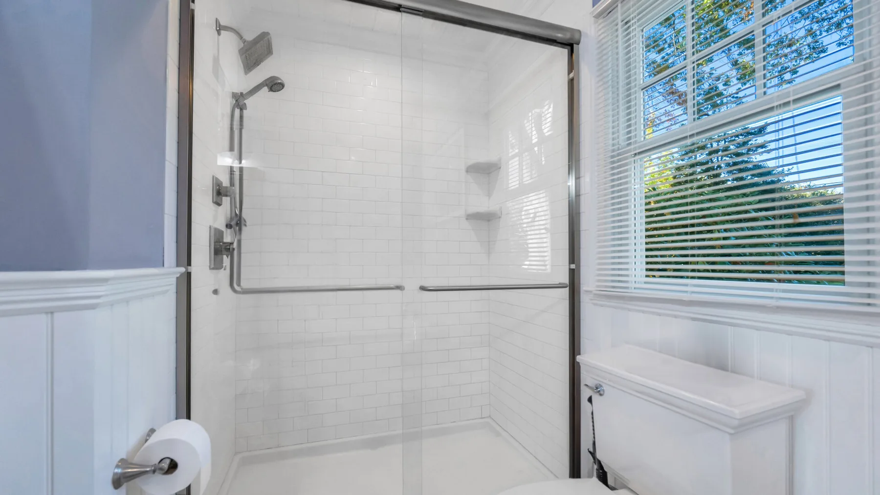 A bright bathroom with a tiled shower and a window with blinds.
