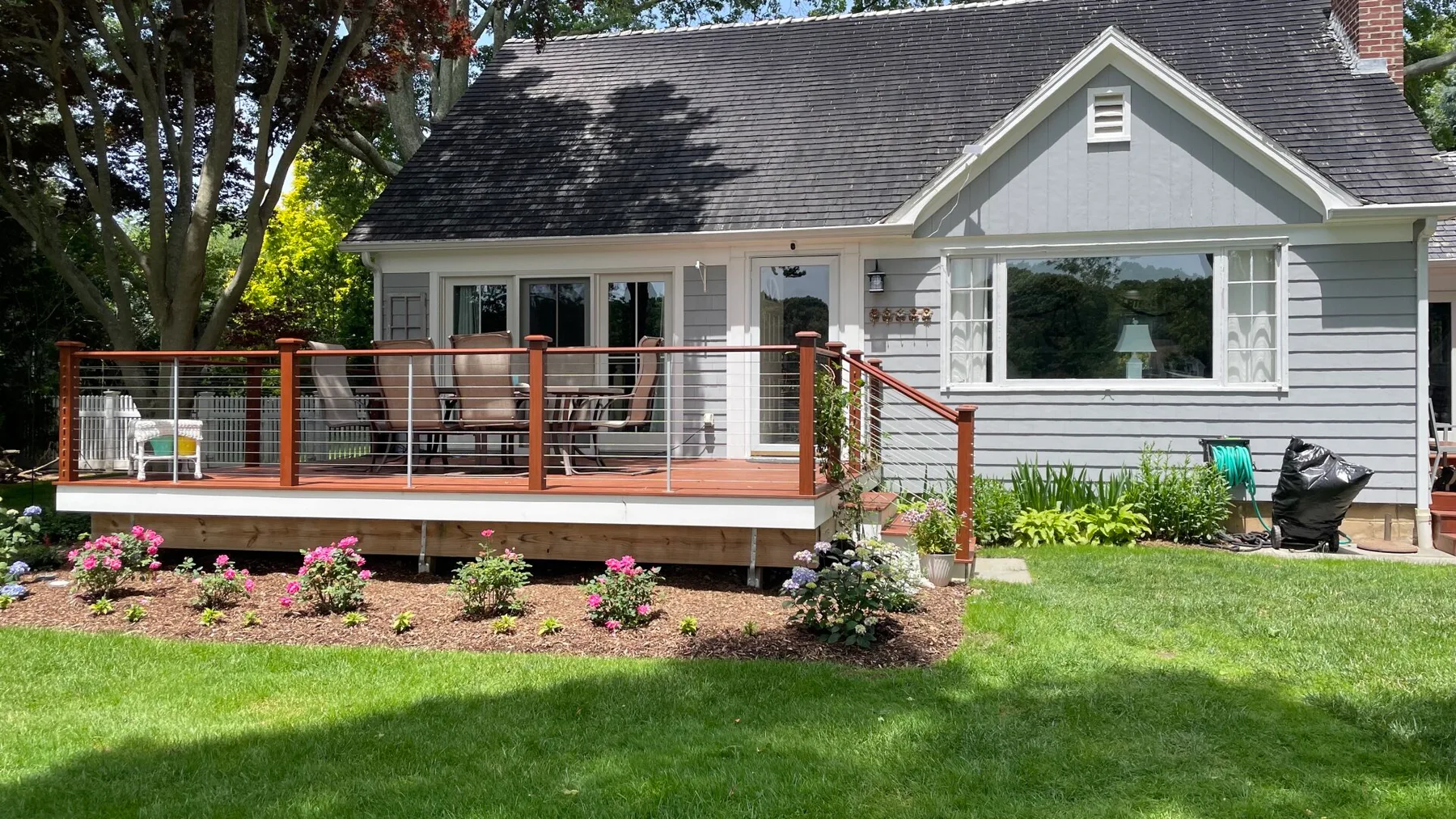 A gray house with a raised wooden deck overlooks a green lawn and garden.