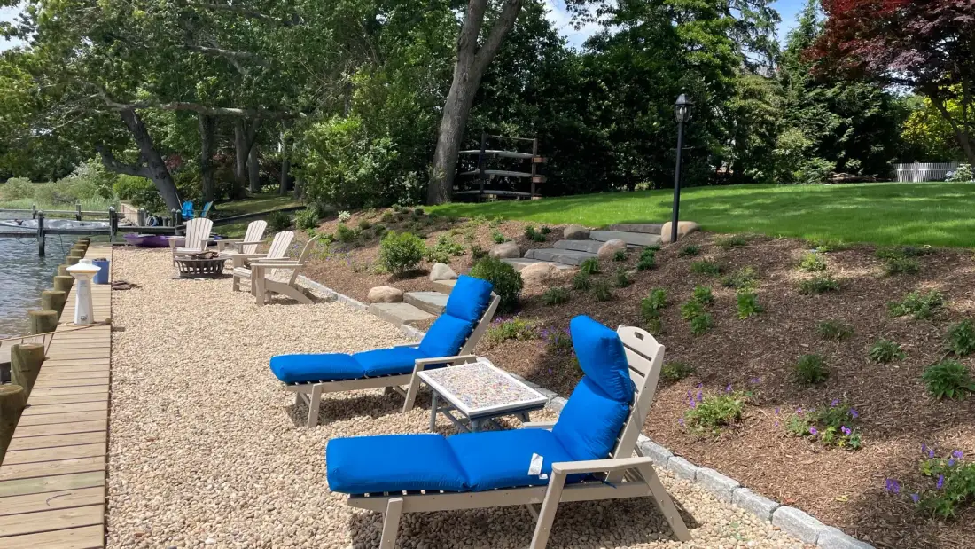 A landscaped lakeside seating area with blue lounge chairs beside a wooden walkway.