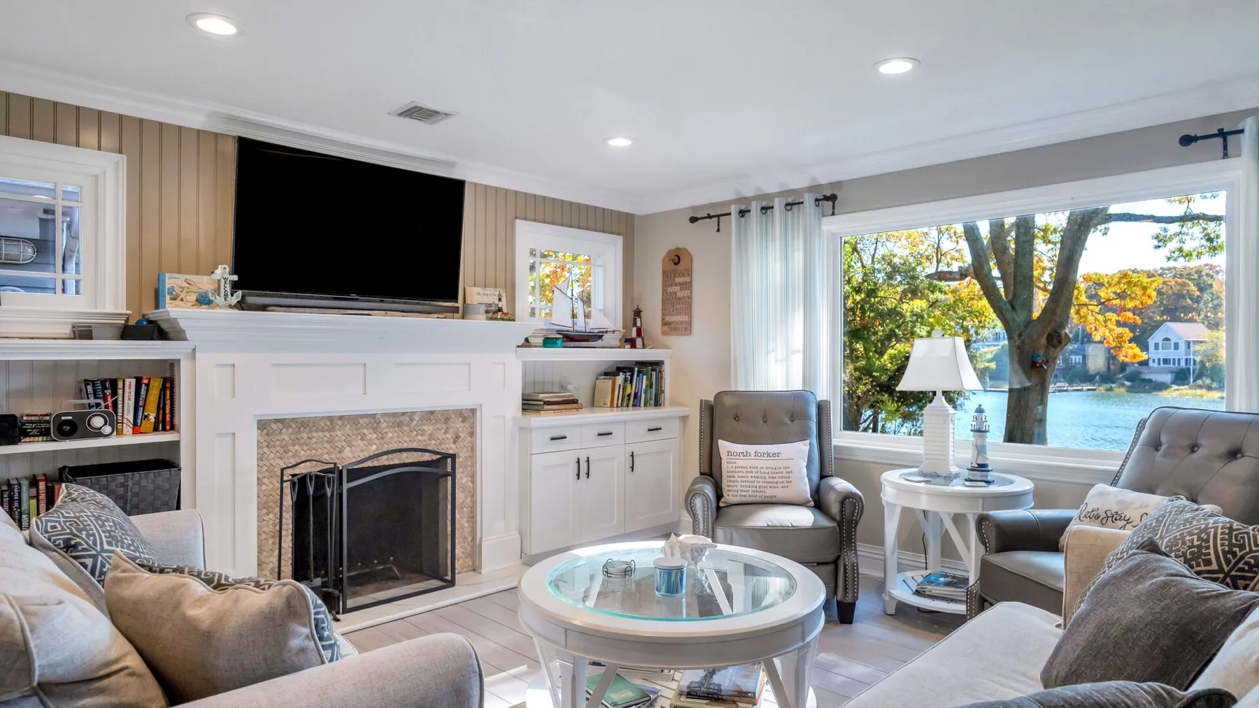 A bright living room with a fireplace, seating, and large windows overlooking trees and water.