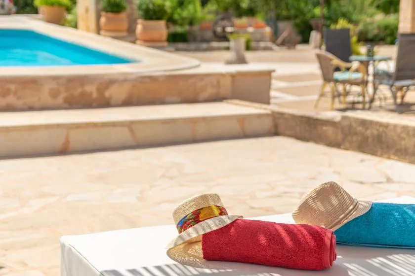 Un patio junto a la piscina con una tumbona a rayas y una toalla roja bajo un sombrero.