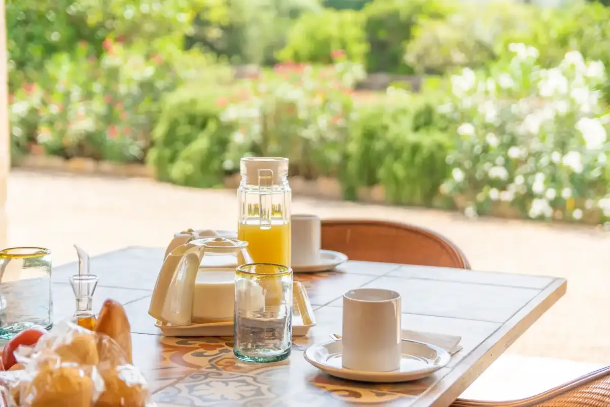 Una mesa al aire libre está preparada con tazas, una jarra de vidrio y alimentos para el desayuno.