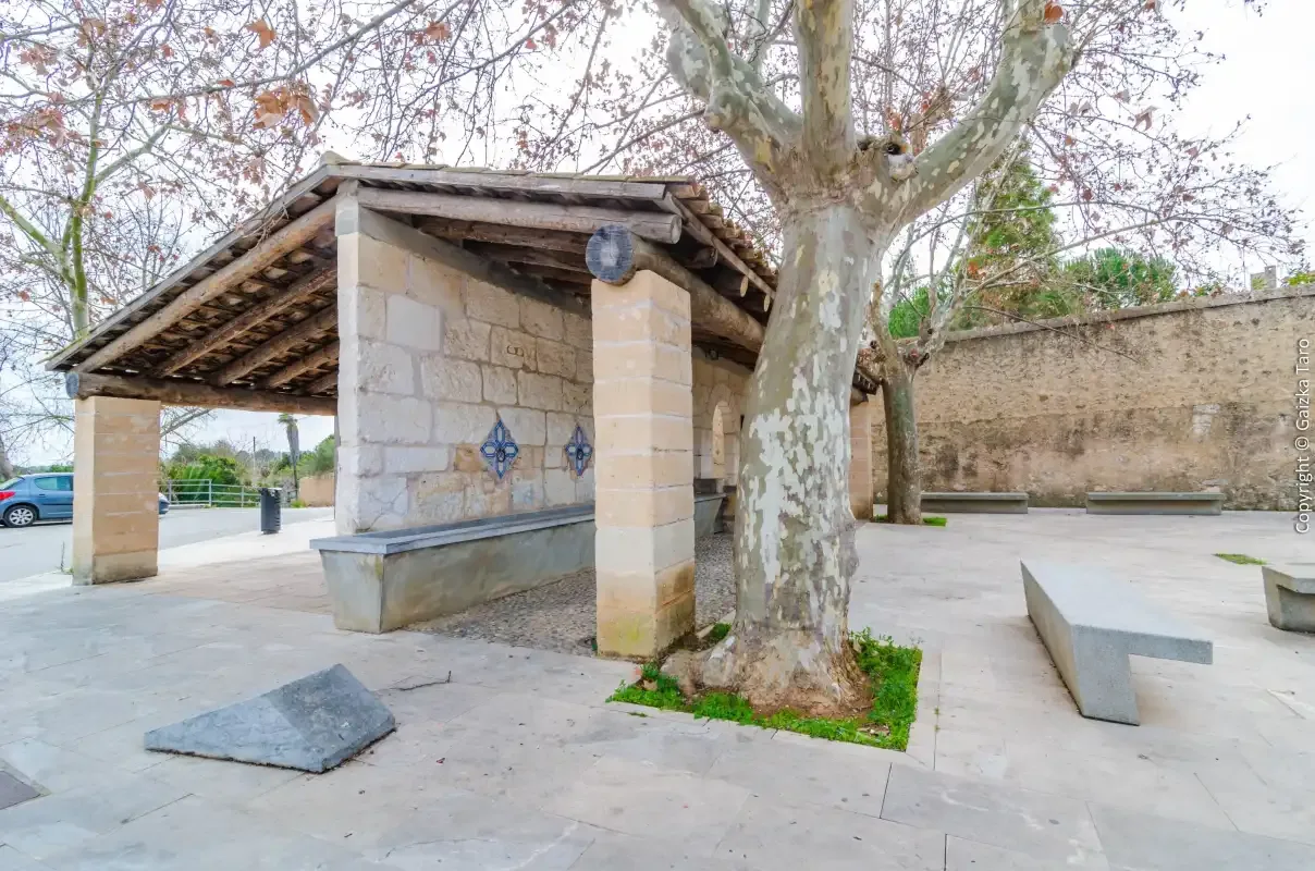 Un refugio de piedra con una fuente se encuentra en un patio pavimentado bajo un gran árbol.