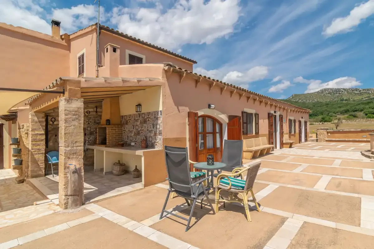 Un patio soleado junto a una casa color melocotón, con asientos al aire libre y una zona de cocina cubierta.
