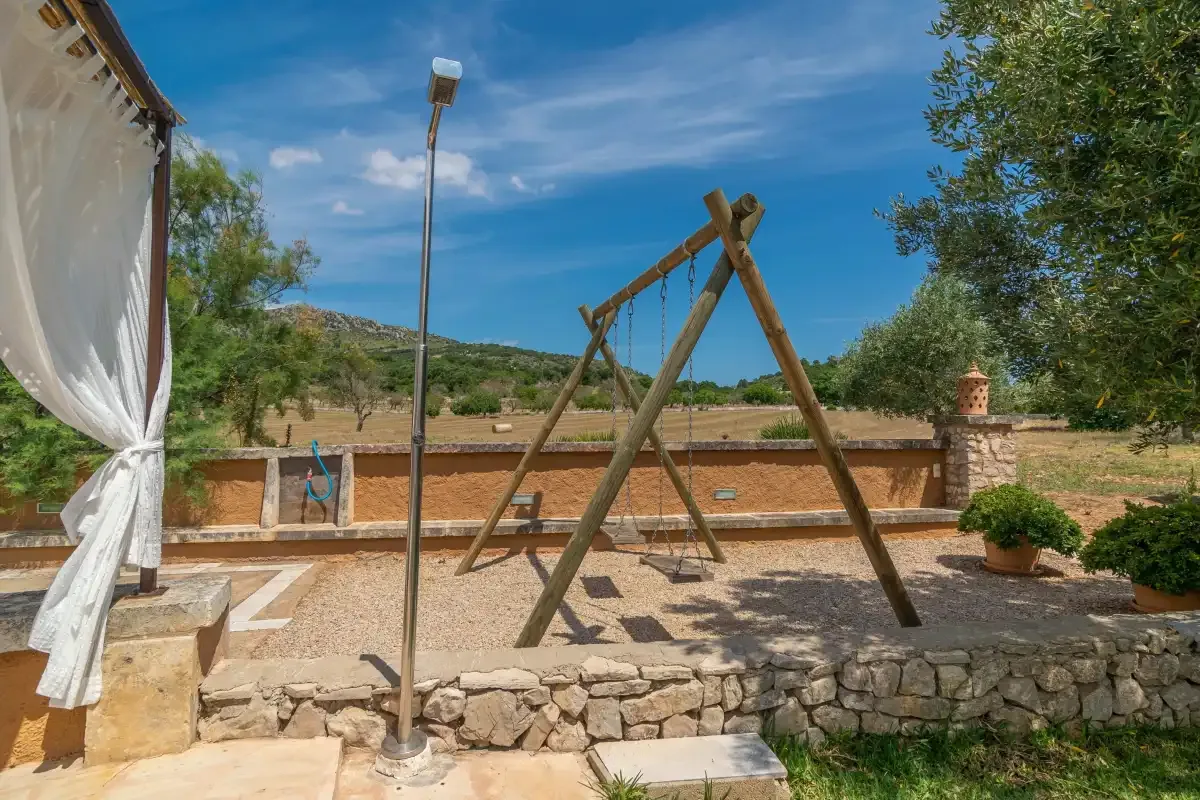 Un columpio de madera está en un patio soleado junto a un muro bajo de piedra.
