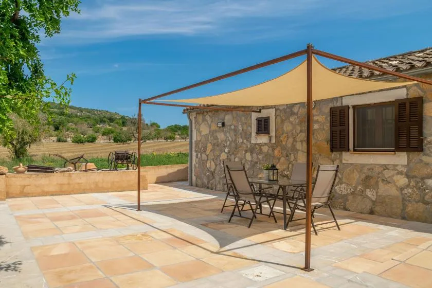 Patio de una casa de piedra con asientos al aire libre bajo una vela de sombra triangular.