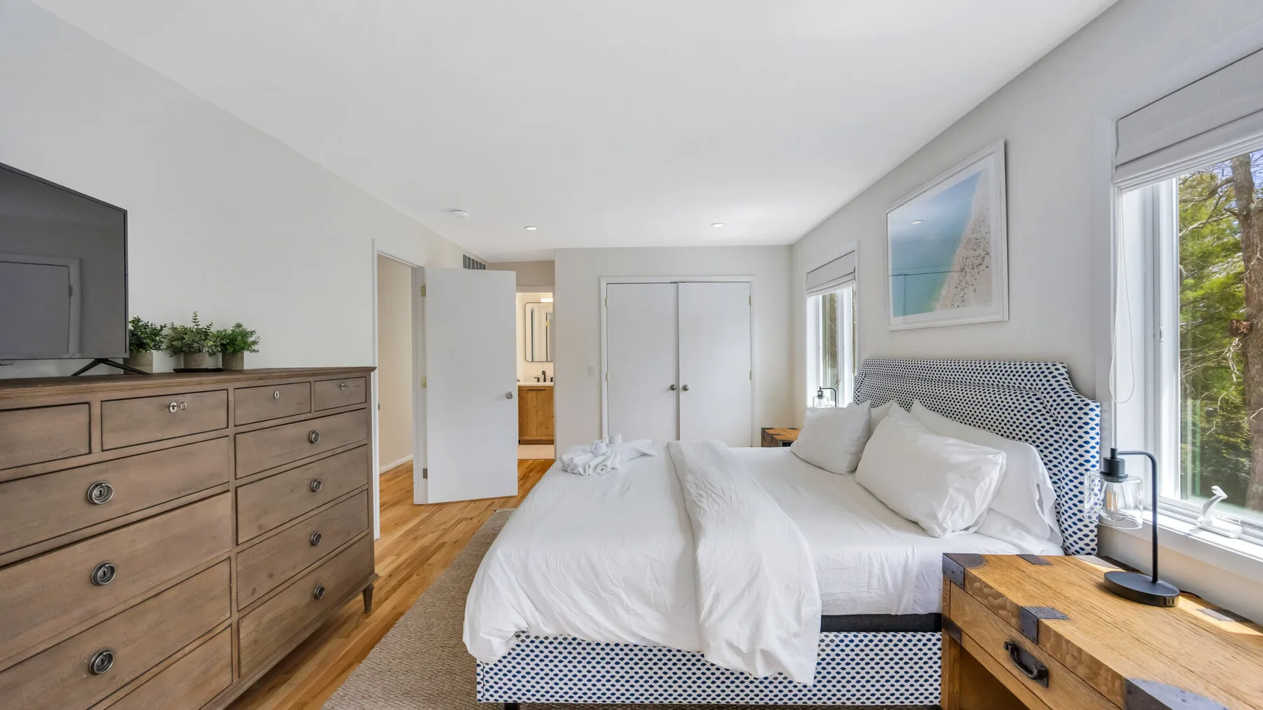 A bright bedroom with a bed, wooden dresser, and large windows.