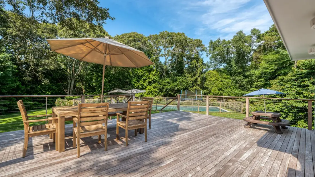 An outdoor wooden deck has dining seating, umbrellas, and trees around it.