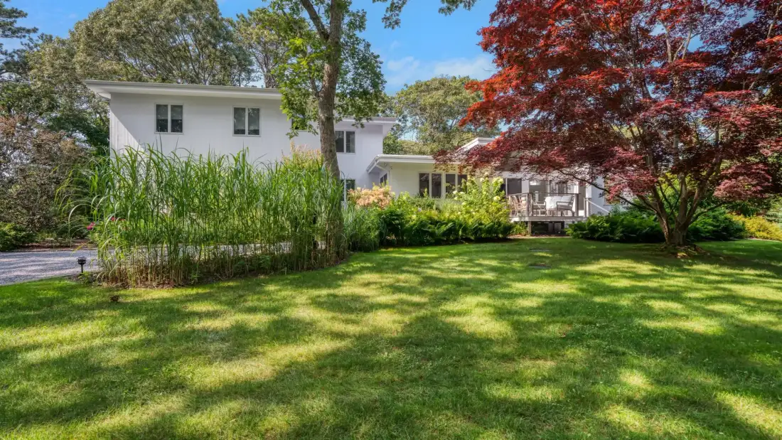 A white house stands behind a green lawn, with leafy trees and garden plants.