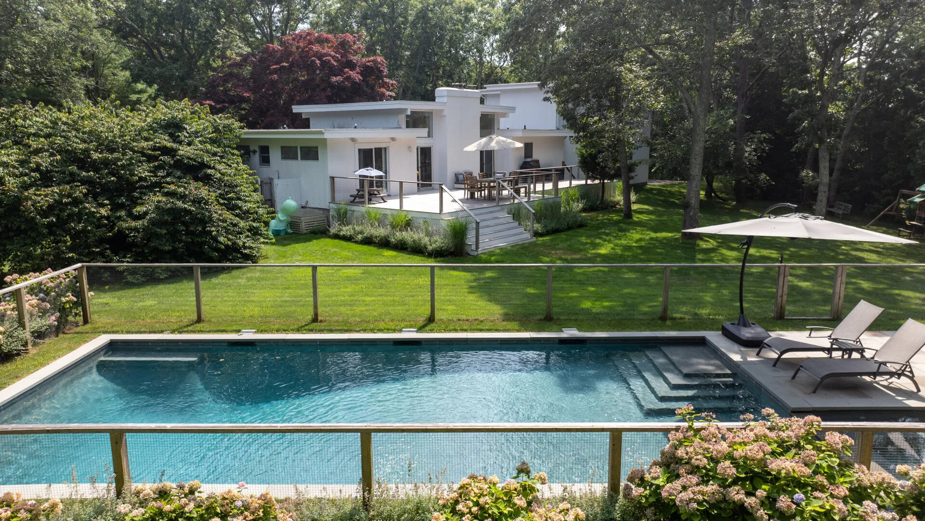 A swimming pool sits in front of a modern house amid green landscaping.