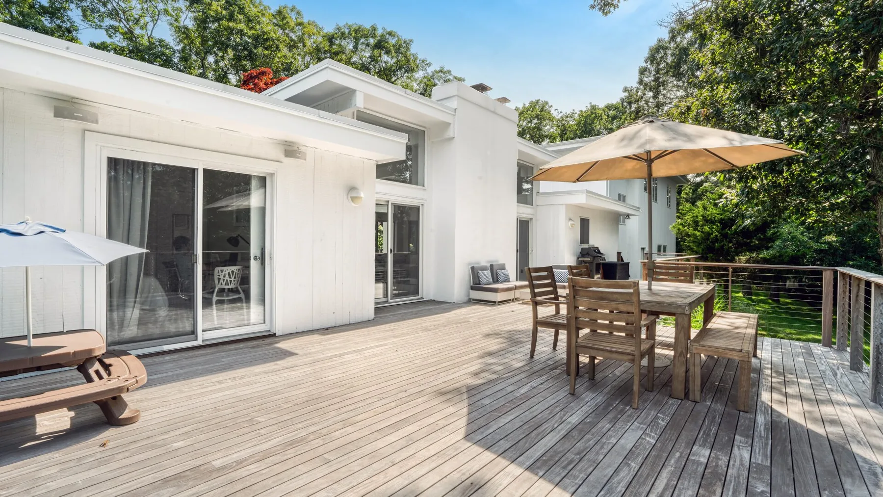 A wooden deck beside a white house has outdoor seating beneath a large umbrella.