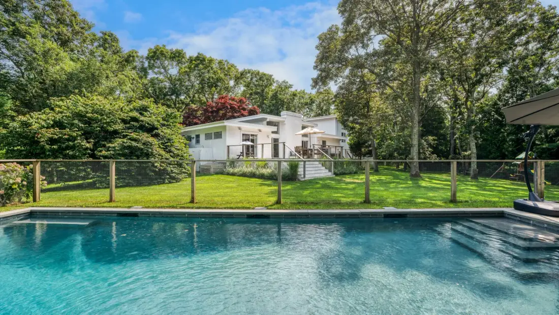 An outdoor swimming pool sits in front of a white house amid trees.