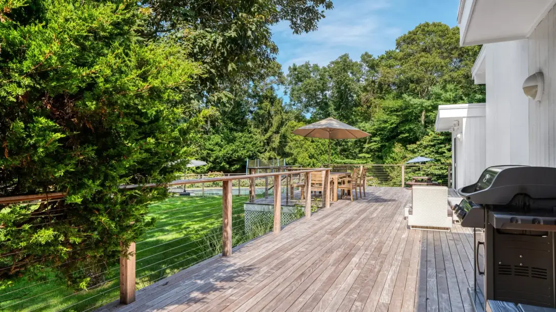 A wooden deck beside a house overlooks a leafy garden with outdoor seating.