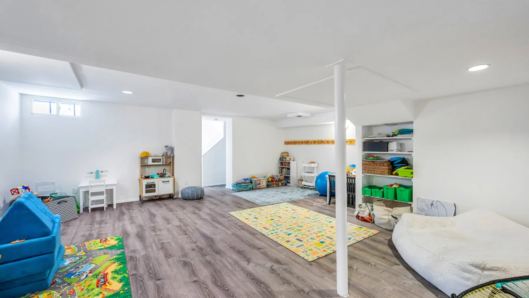 A bright playroom has colorful floor mats, children's toys, and open storage shelves.