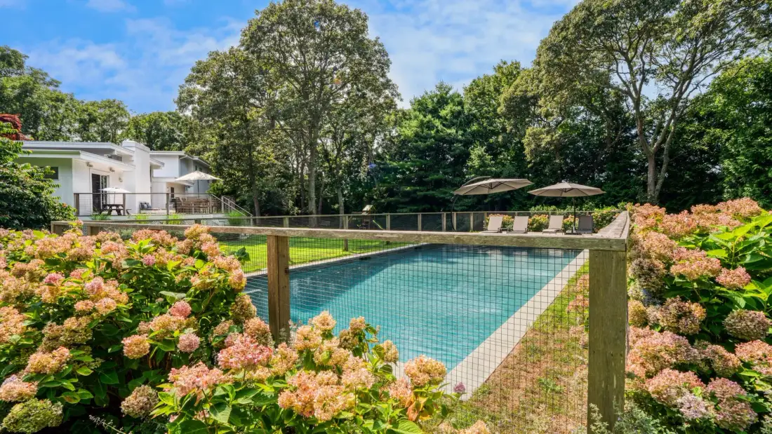An outdoor swimming pool sits behind a wooden fence, surrounded by flowering shrubs and trees.