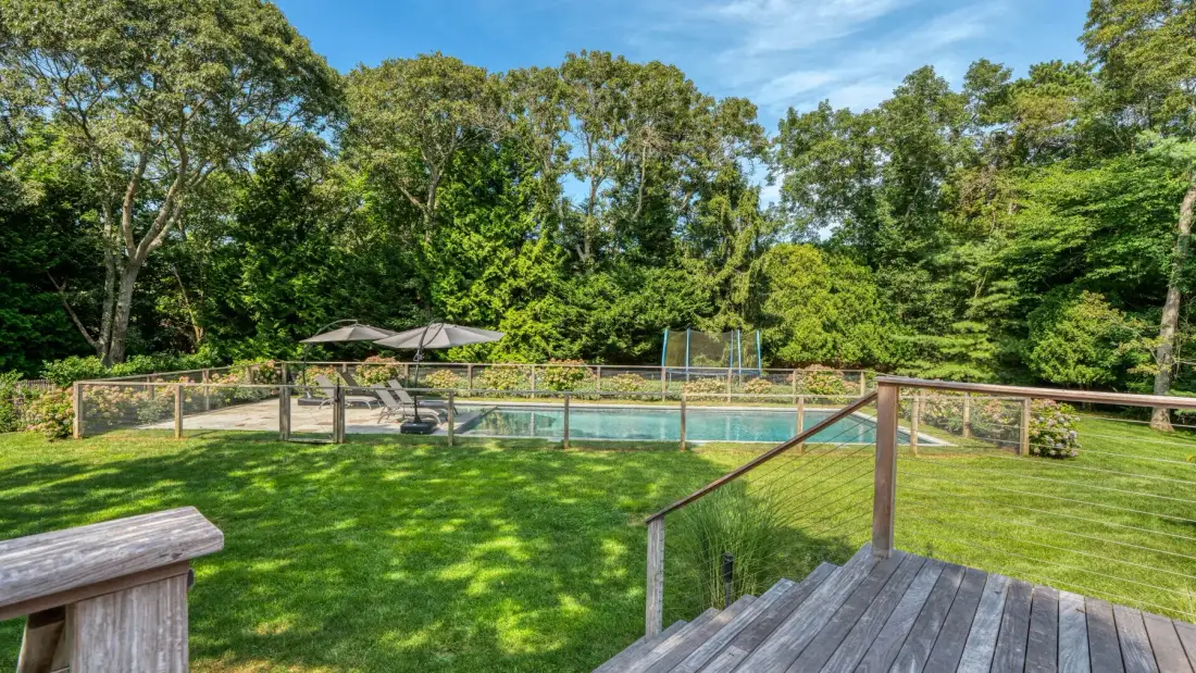 A fenced outdoor swimming pool sits beside a grassy lawn and wooded surroundings.