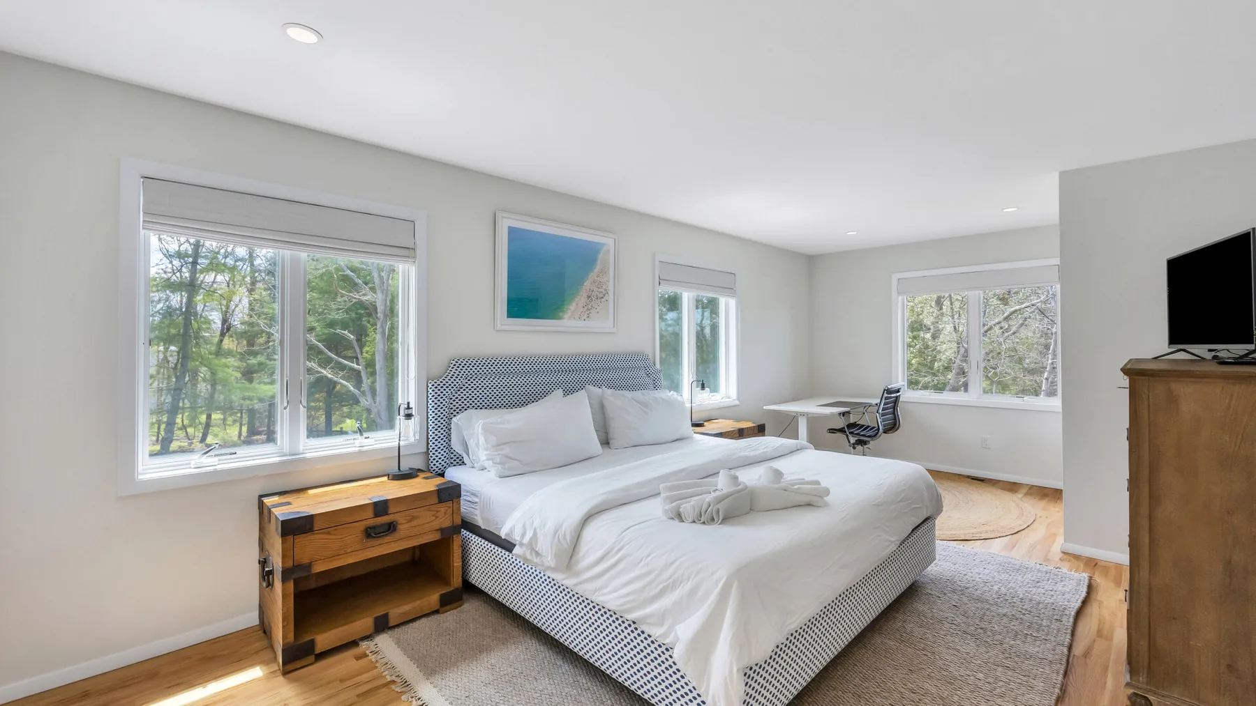 A bright bedroom with a bed, bedside table, and windows overlooking trees.