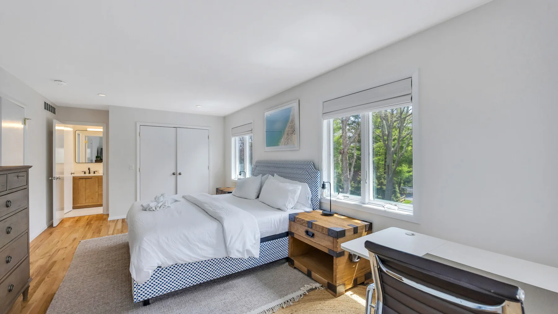 A bright bedroom with a bed, a desk, and windows overlooking trees.
