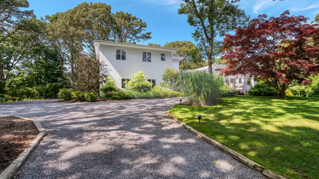 A paved driveway leads toward a white house among trees and landscaping.