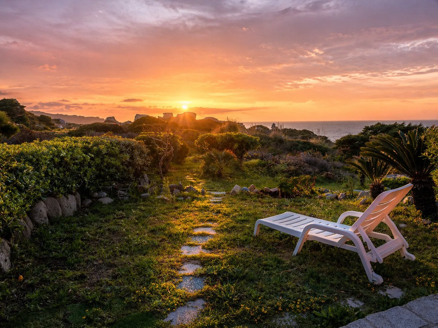 A garden path leads through coastal greenery toward the ocean at sunset.