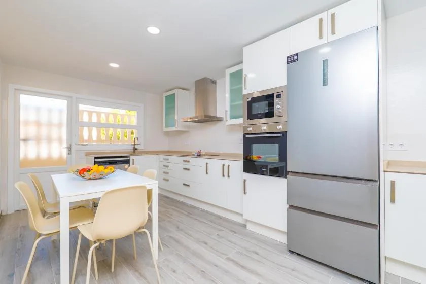 Una cocina luminosa con gabinetes blancos, una mesa y un refrigerador grande.