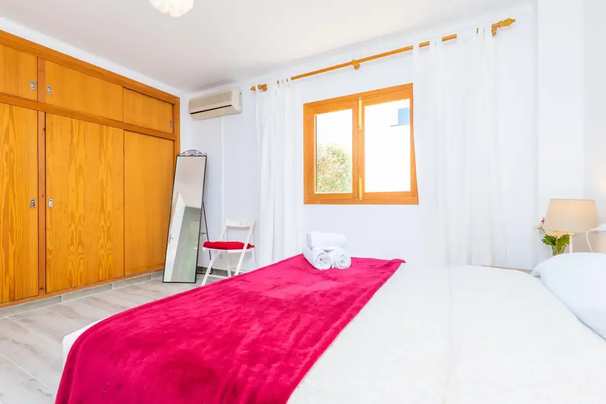 Un dormitorio luminoso con una cama, una manta roja y un armario de madera.