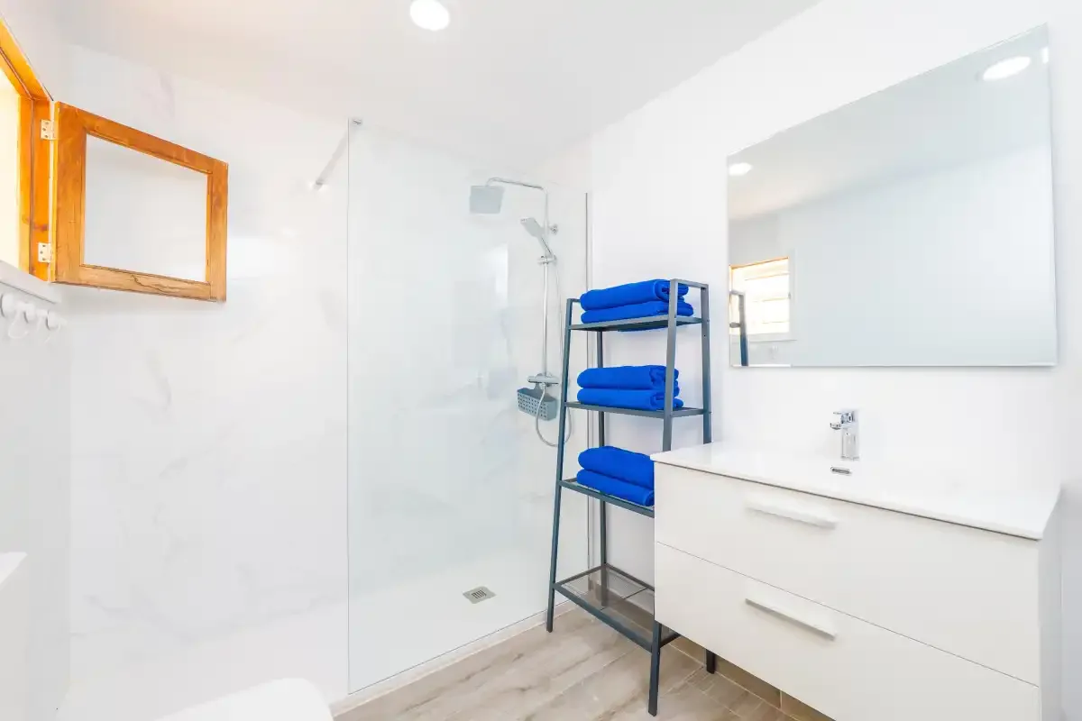 Baño luminoso con ducha, lavabo y toallas azules en una estantería.