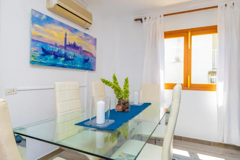 Un comedor luminoso con una mesa de vidrio, asientos blancos y una ventana soleada.