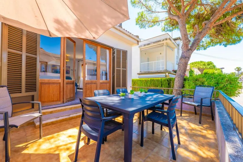 An outdoor patio with a dining table and seating beside glass doors and greenery.