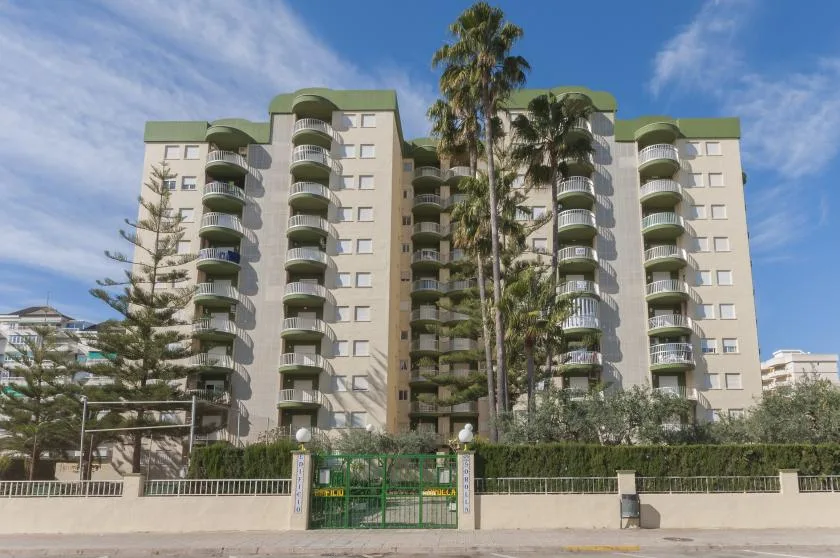 Un edificio alto de apartamentos con balcones curvos se alza tras una entrada verde.