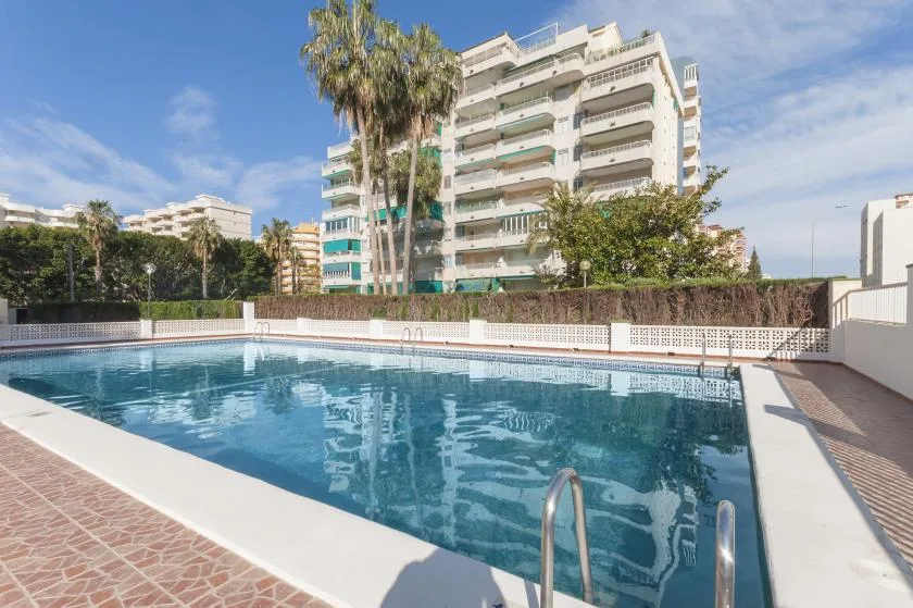 An outdoor swimming pool sits beside a tall residential building under a blue sky.