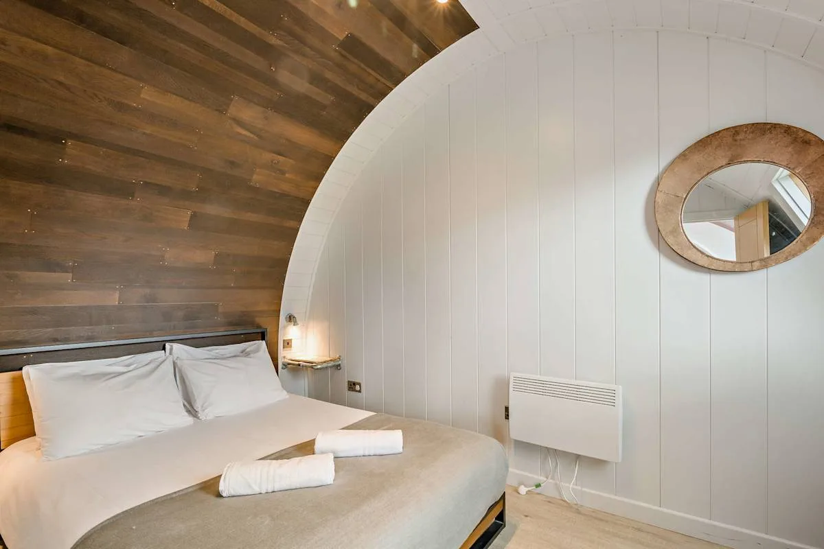 A bedroom with a bed, white paneled walls, and a round wall mirror.
