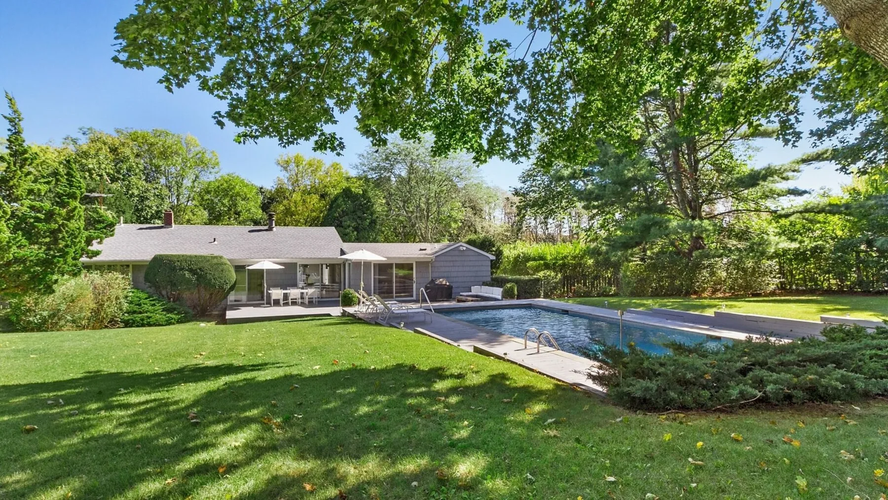 Un patio trasero con piscina, césped y una casa bajo árboles frondosos.