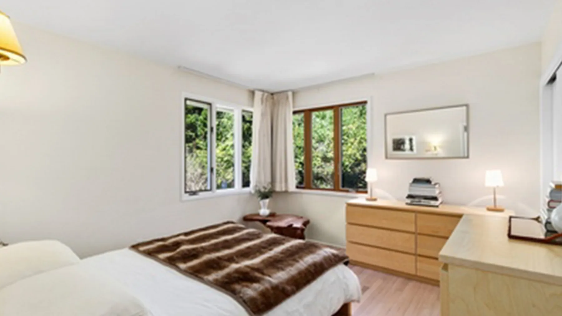 Un dormitorio luminoso con una cama junto a grandes ventanas y una cómoda de madera.
