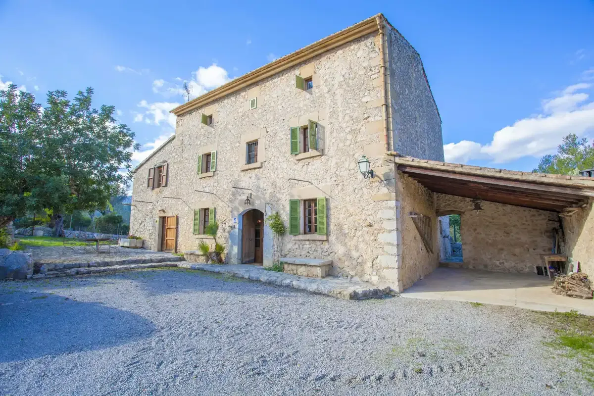 A stone building with green shutters stands beside a covered parking area.