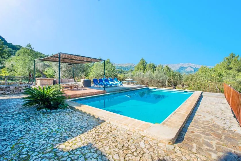 Una piscina al aire libre está junto a un patio de piedra, con tumbonas azules bajo una pérgola sombreada.