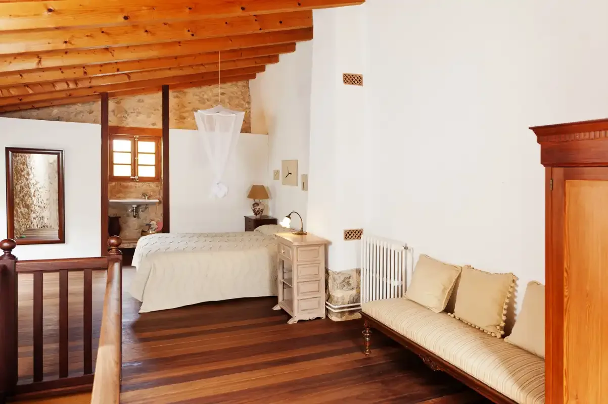 A bedroom with a bed, a cushioned seating area, and exposed wooden ceiling beams.