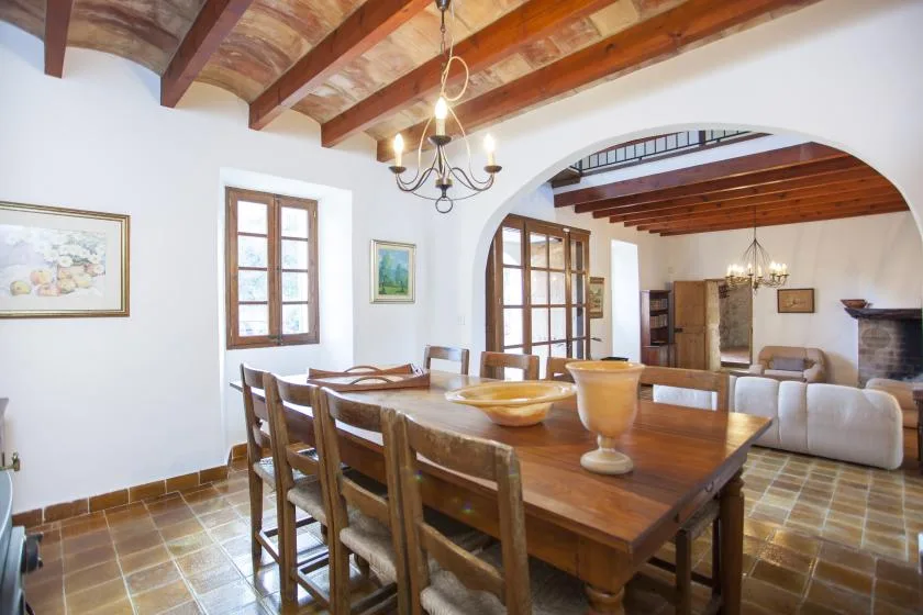 A wooden dining table and seating sit beneath exposed ceiling beams in a bright room.